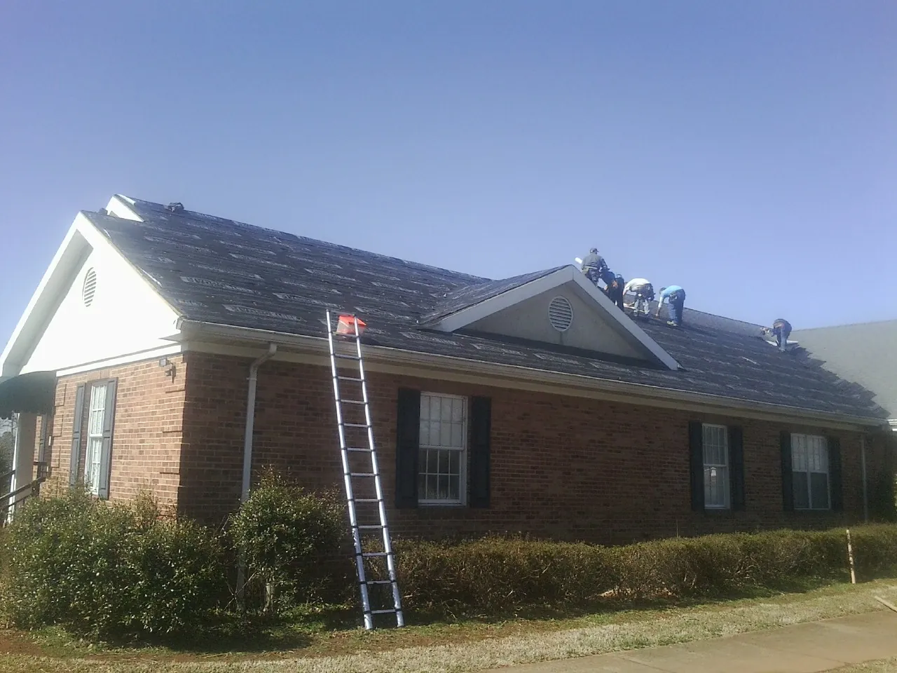 Crew tearing shingles off a brick ranch roof with a ladder set — Emergency Roof Repair work by our crew in Ulen