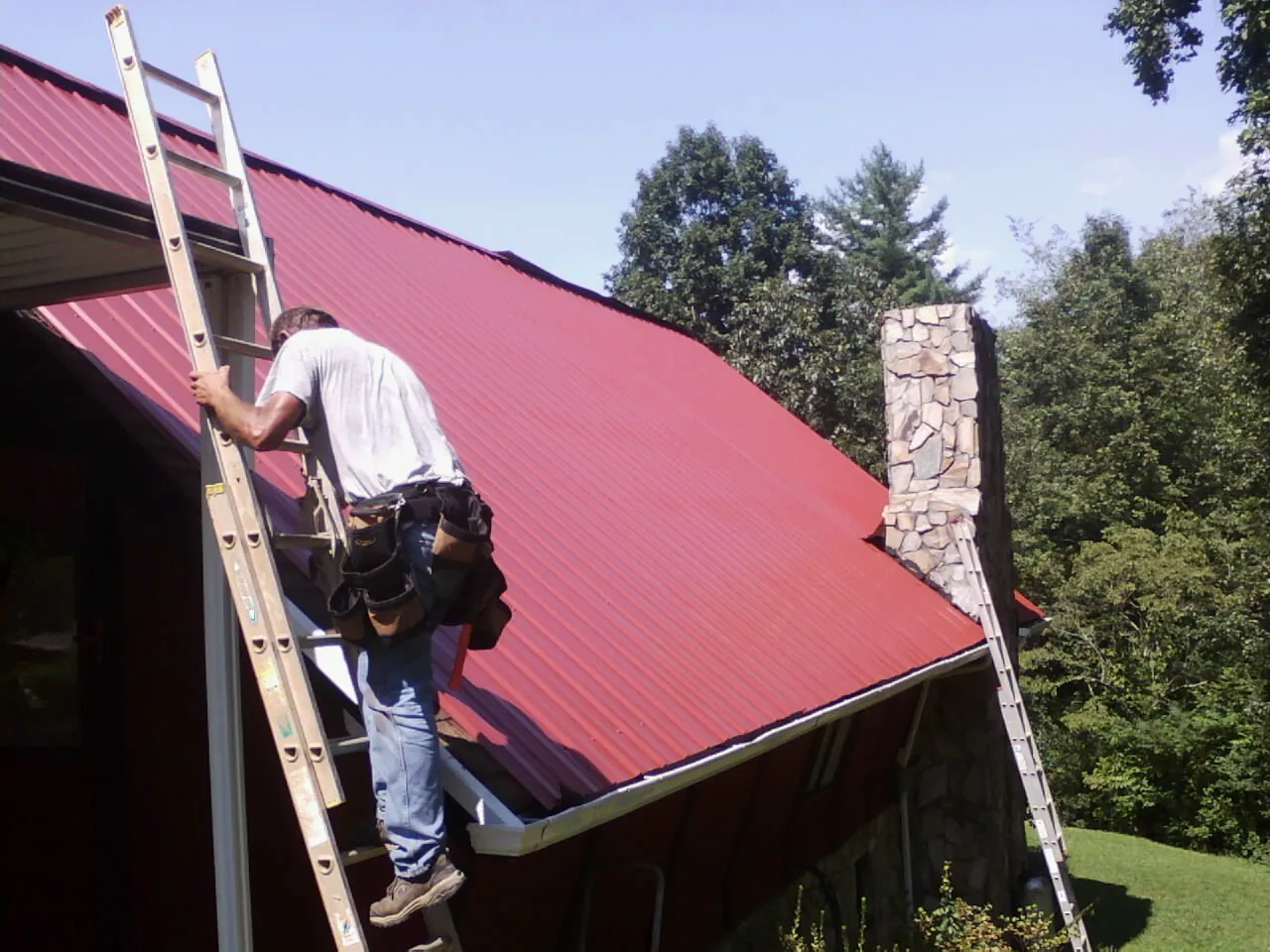 Roofer on a ladder beside a red metal roof and stone chimney — Soffit & Fascia Repair by Roofing Brothers in Ulen